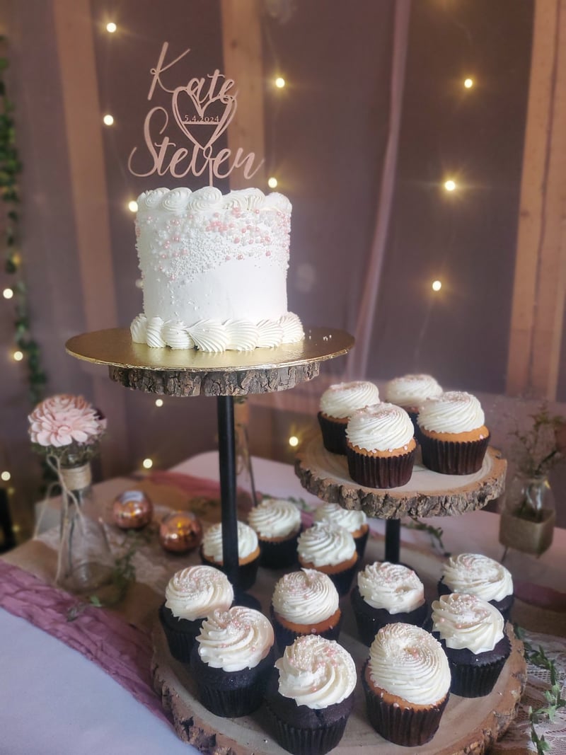 A wedding cake adorned with pink and white sprinkles, placed on a rustic cake stand. Surrounding it are cupcakes with white frosting, on a tiered wooden display, in a decorated venue with warm lights and floral arrangements.