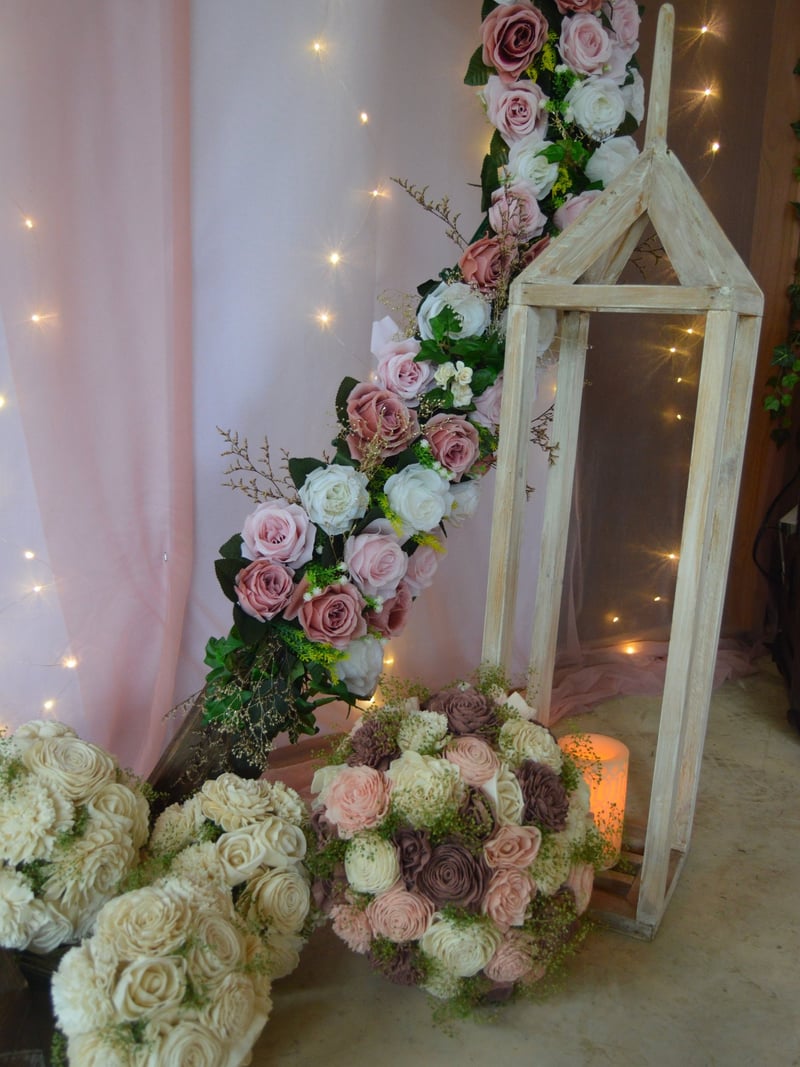 Wedding arrangement of pink, white, and purple roses with greenery displayed with a wooden frame and pink fabric backdrop, decorated with fairy lights.