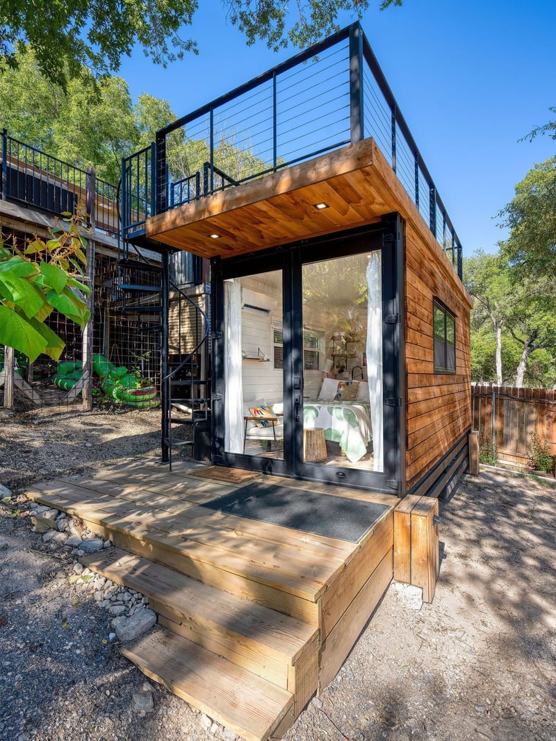 A modern tiny home with a wooden exterior and black trim, featuring large glass doors and a rooftop deck with a black metal railing. A spiral staircase leads to the upper level, and wooden steps lead to the entrance. The interior is visible through t