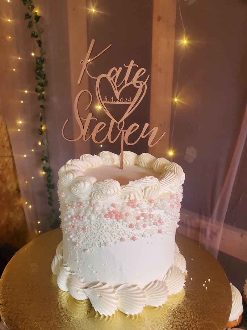 A white wedding cake decorated with pink and white sprinkles and piped cream, topped with a wooden cake topper with yellow fairy lights and greenery in the background.