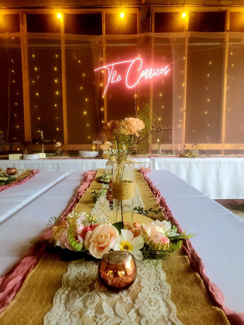 Decorated banquet wedding table with floral centerpiece, candle, and pink fabric runner, set against a backdrop of string lights and a neon sign