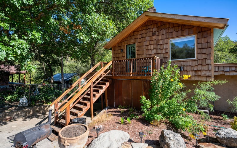 A rustic cabin with wood shingle siding and a small balcony, accessed by a wooden staircase. The surrounding area features lush plants, large rocks, and a charcoal grill on a concrete patio, shaded by tall trees.