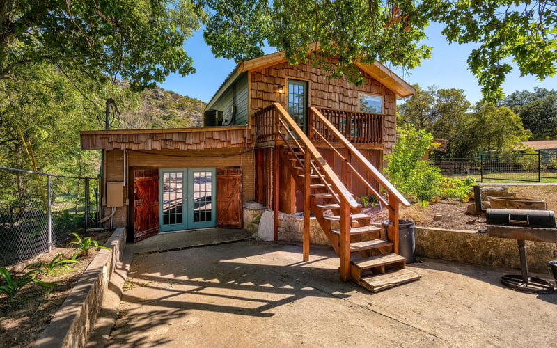 A rustic two-story house with wood shingles and green siding. A wooden staircase leads to an upper-level balcony, while French doors with wooden shutters open to the lower level. A charcoal grill sits on the concrete patio, surrounded by trees and gr