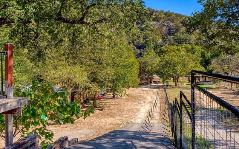 A sloped pavement road with a black railing leading down to a rustic campground with small wooden cabins, trees, and open space. A dirt road extends into the distance, with rolling hills and greenery in the background.