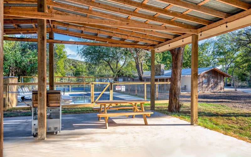 A covered outdoor pavilion with a wooden picnic table and stainless steel grill, overlooking a fenced sports court and a rustic house among trees.