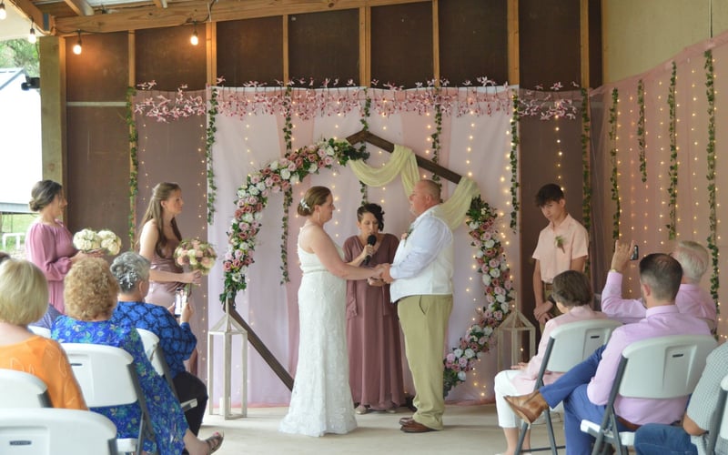 A wedding ceremony taking place in an indoor venue with a decorated backdrop featuring pink and white flowers, fairy lights, and greenery. The bride and groom are holding hands in front of an officiant, with bridesmaids and groomsmen standing nearby.