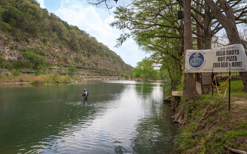 A person standing in shallow water while fishing by a riverside lined with trees and a rocky hillside.