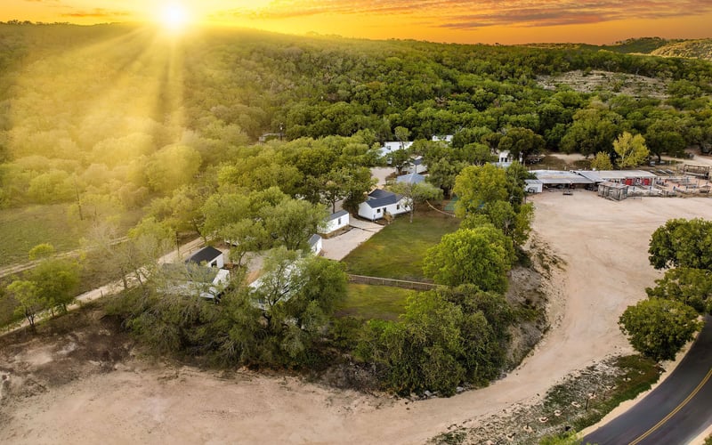 Aerial view of a scenic New Braunfels Vacation rental property at sunset, featuring modern white cottages surrounded by lush greenery, rolling hills, and a winding road.
