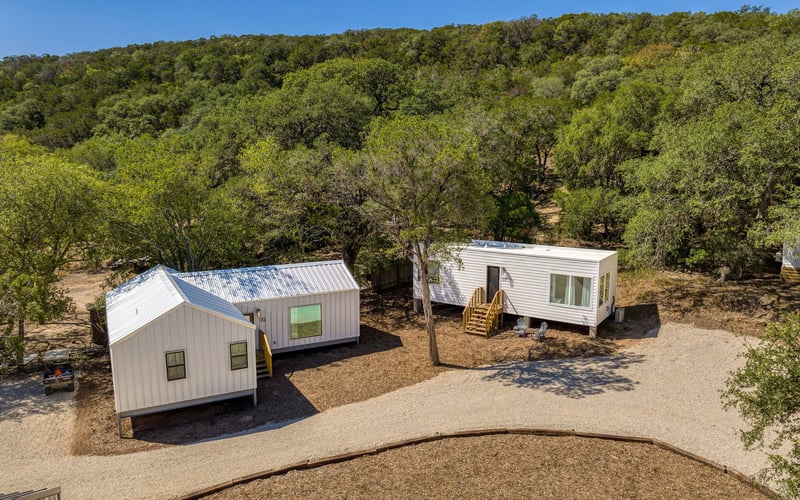 Scenic countryside vacation rental with modern white cabins, wooden stairs, and gravel pathways, surrounded by lush greenery and rolling hills.