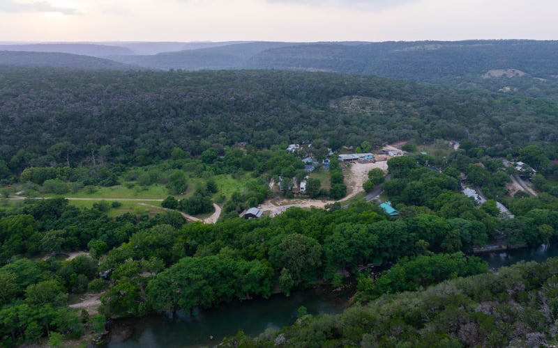 Aerial view of a lush green landscape with a river running through it, scattered houses and buildings, and mountains in the background.