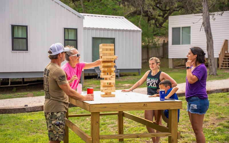 People playing giant Jenga outdoors on a wooden table, with cottages in the background in New Braunfels, Texas Hill Country.