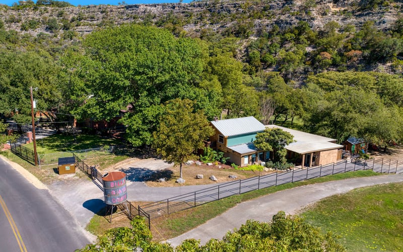 Rustic cabin-style New Braunfels Vacation Rental with a metal roof, covered patio, and gated driveway, surrounded by lush trees and scenic hillside views.
