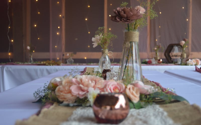Decorated wedding table with vases of pink and brown flowers, surrounded by string lights and a rustic interior background, for a wedding or special event.