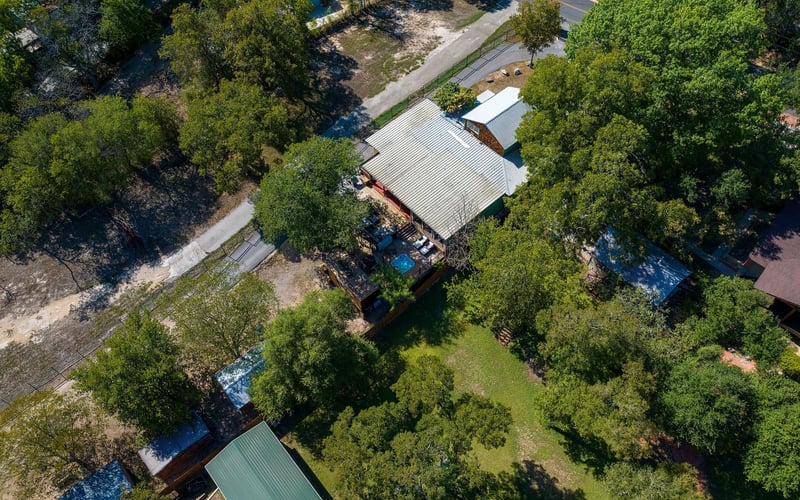 A secluded New Braunfels Vacation Rental property with a metal-roofed main house, outdoor decks, a hot tub, and lush tree coverage along a quiet road.