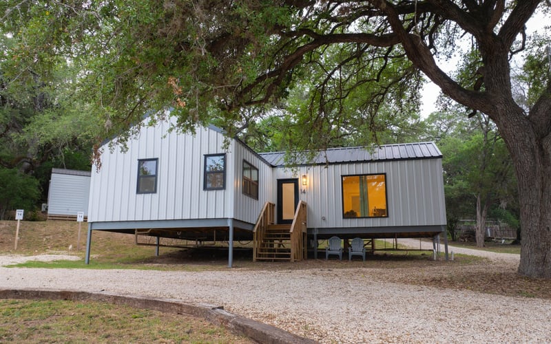 A modern tiny house with white metal siding and a metal roof, elevated on stilts, with small staircase leading to the front door, surrounded by large trees and a gravel driveway.