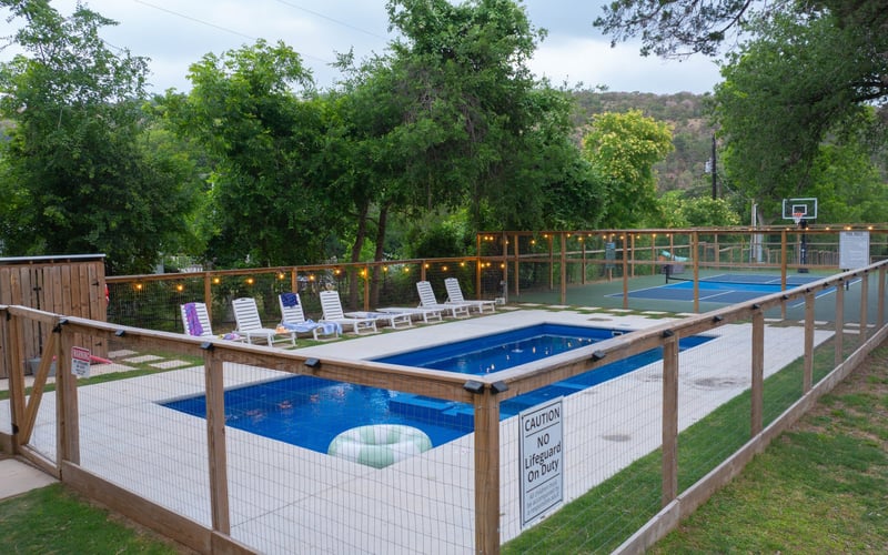 A fenced backyard with a small pool, a hot tub, lounge chairs, and a nearby basketball court surrounded by trees.