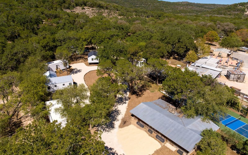 Secluded vacation property with modern tiny homes, a large covered pavilion, winding gravel paths, and a tennis court, surrounded by rolling hills and dense trees.
