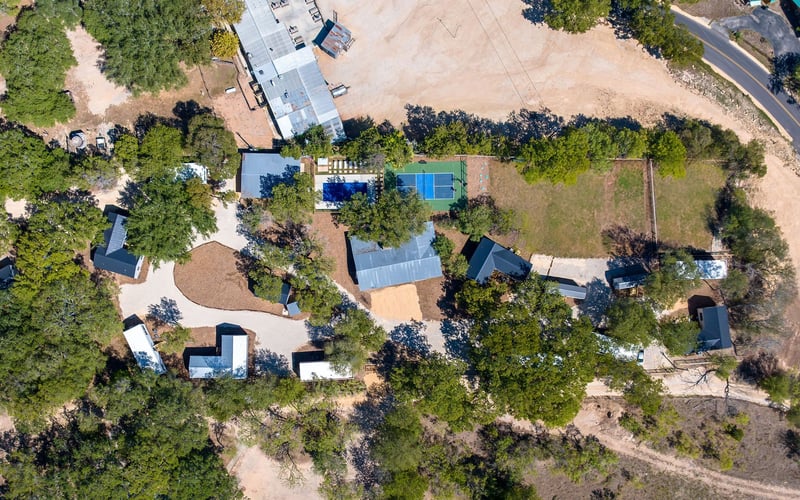 Aerial view of a countryside New Braunfels Vacation Rental property featuring multiple cabins, a pickleball court, open green spaces, and a main lodge surrounded by trees.