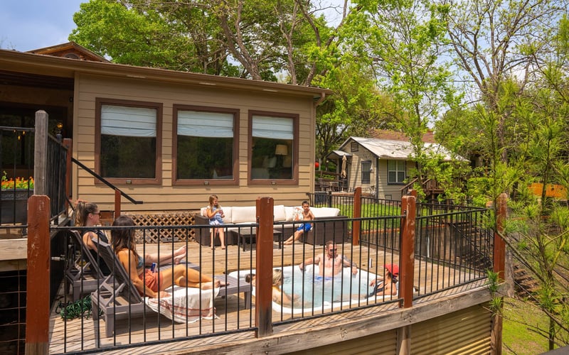 People relaxing on a wooden deck with a hot tub and seating area, surrounded by trees and adjacent to houses, during daytime.