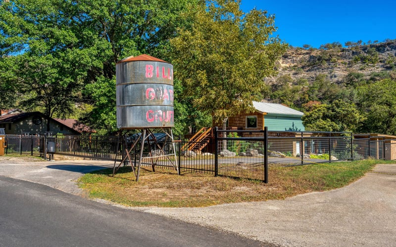 A rustic property enclosed by a black metal fence, featuring a weathered water tower with 'Billy Goat Gruff' painted in red. A wooden cabin with a green upper level and a staircase is nestled among trees, with a rocky hillside in the background.