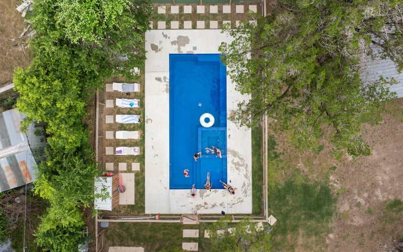 A top-down view of a Fernweh swimming pool with four people sitting on the edge and two children playing in the pool. Surrounding the pool are trees, lounge chairs, and a concrete patio.