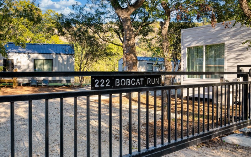A black metal gate with a sign reading '222 Bobcat Run' opens to a gravel driveway lined with modern cabins, trees, and outdoor seating.
