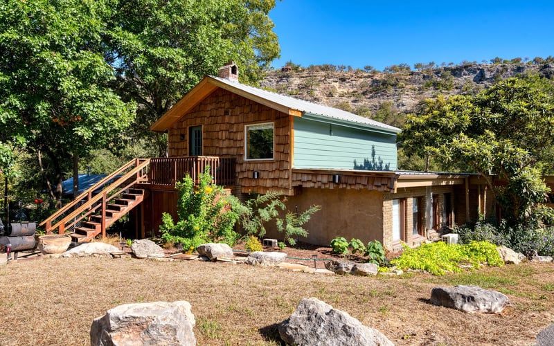 A rustic two-story house with wood shingles and green siding, set against a hillside. A wooden staircase leads to an upper-level deck, surrounded by lush greenery, large rocks, and a landscaped yard.