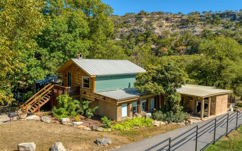 Rustic two-story cabin with wood siding, green accents, a covered patio, and outdoor stairs, set against a scenic hillside with lush trees.