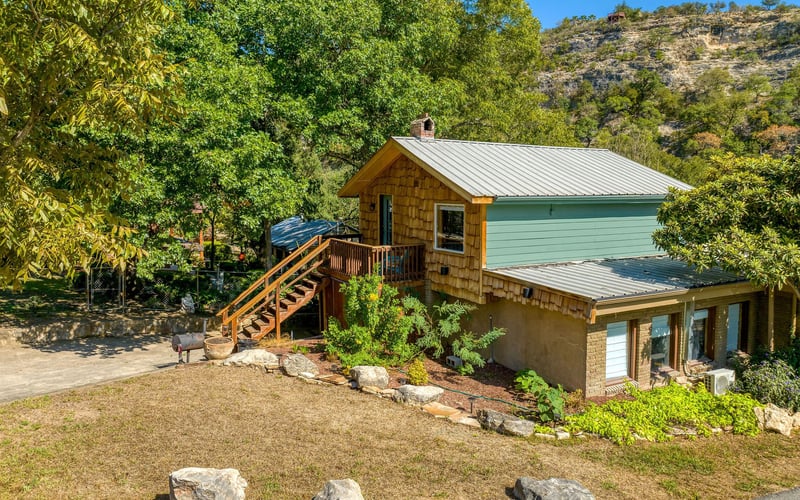 Rustic two-story cabin with wood shingles, green siding, a wraparound deck, and outdoor stairs, nestled in a scenic hillside with lush trees.
