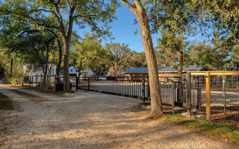 A gated entrance with a black metal and wood fence along a gravel driveway, surrounded by tall trees and rustic buildings in the background.
