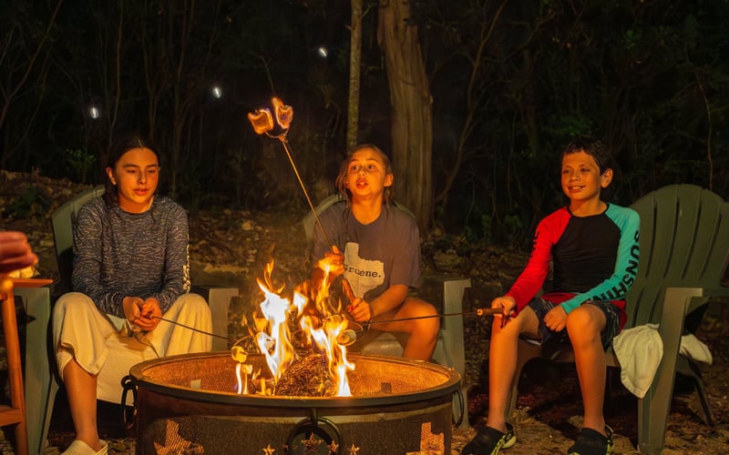 Three kids sitting around a campfire at night, roasting marshmallows on sticks, in a wooded outdoor setting.