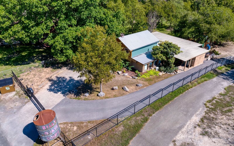 Rustic cabin-style home with a metal roof, covered patio, and gated driveway, surrounded by lush trees and scenic hillside views.