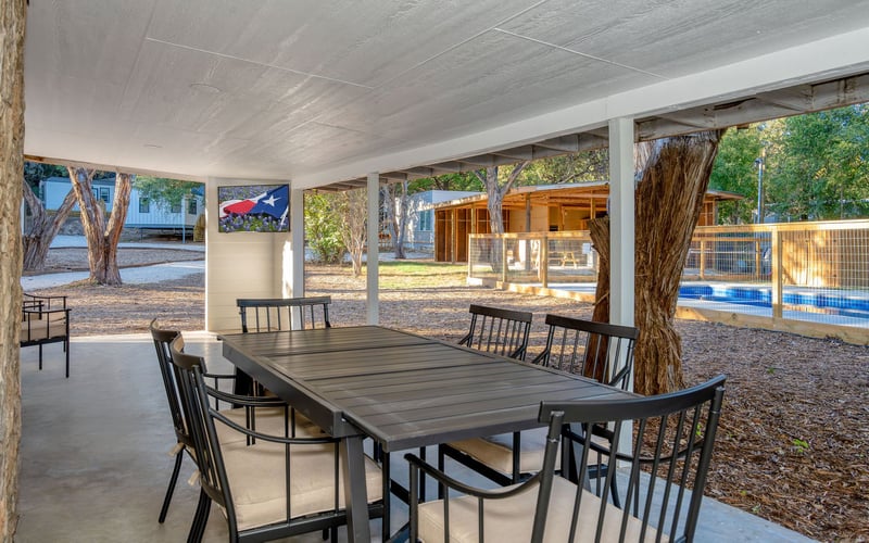 Covered outdoor dining area with a black metal table and chairs, overlooking trees, a wooden structure, and a sports court with a blue surface. A Texas-themed artwork hangs on a white post.