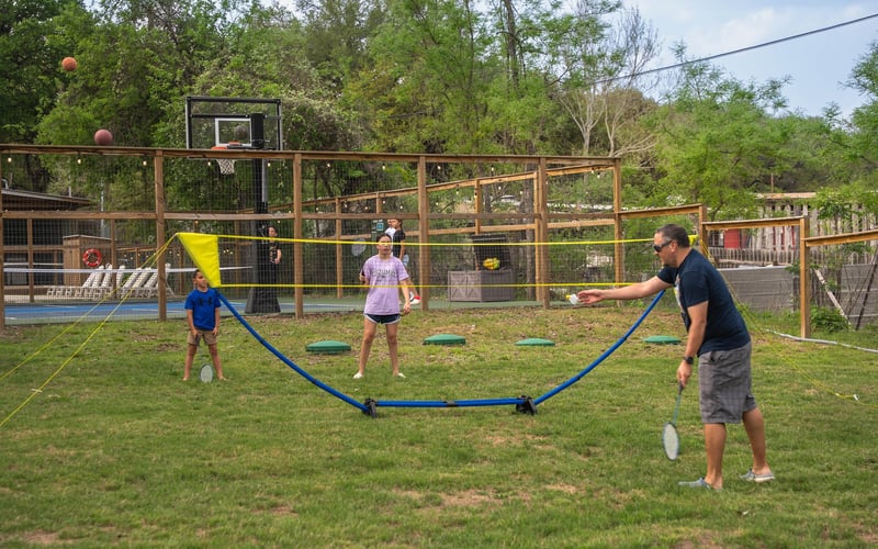 People playing pickleball on a grassy outdoor court with a basketball hoop, surrounded by trees and a wooden fence.