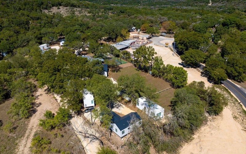Countryside retreat featuring cozy rental cabins, a treehouse-style home, open green spaces, and a nearby marketplace, all set within rolling hills and dense woodlands.