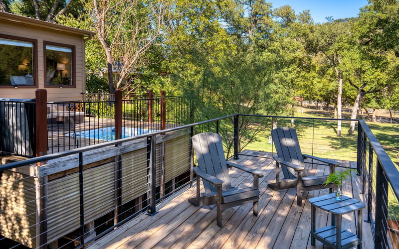 A wooden deck with two black Adirondack chairs and a small side table, overlooking a lush green landscape. A modern cabin with large windows and a pool is visible in the background, enclosed by a black metal railing.