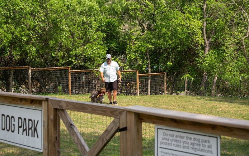 A man walking his dog in a dog park with green trees in the background and a wooden fence. The dog is carrying a yellow toy in its mouth.