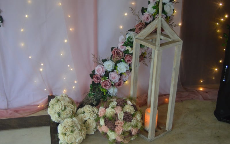 Wedding arrangement of pink and white roses with greenery and fairy lights on a pink fabric backdrop.
