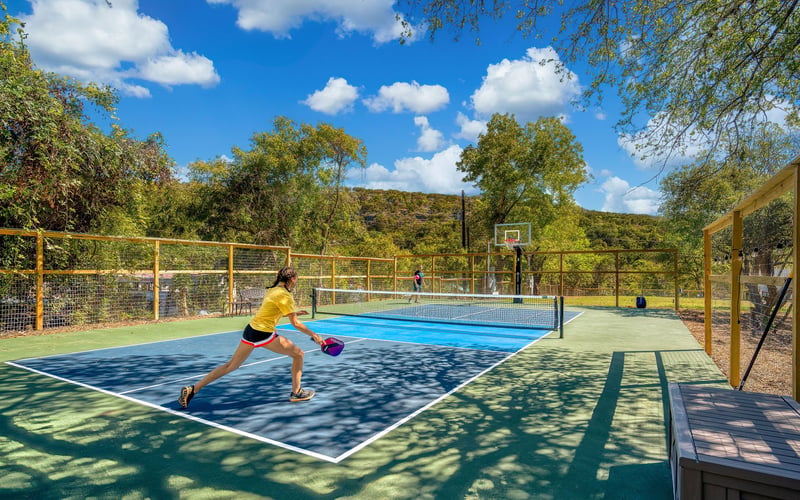 A woman in a yellow shirt plays pickleball on an outdoor court with a blue and green surface, surrounded by a wooden fence. Another player stands on the opposite side near a basketball hoop. Trees and rolling hills fill the background under a bright 