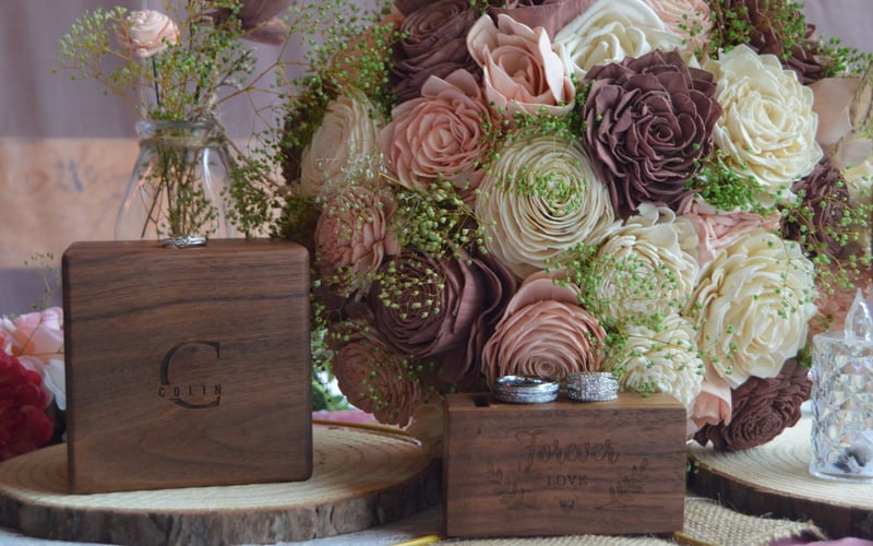 A bouquet of large, ruffled roses in shades of pink, white, and purple, with small green filler flowers, is placed behind two wooden boxes on a wooden slice. The boxes are decorated with engraved text, and there are rings placed on top of one of the 