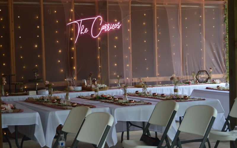 Wedding reception setup with long white tables, floral centerpieces, and pink accents. A pink neon sign reads 'The Carricos,' and the backdrop features string lights and pink sheer fabric.