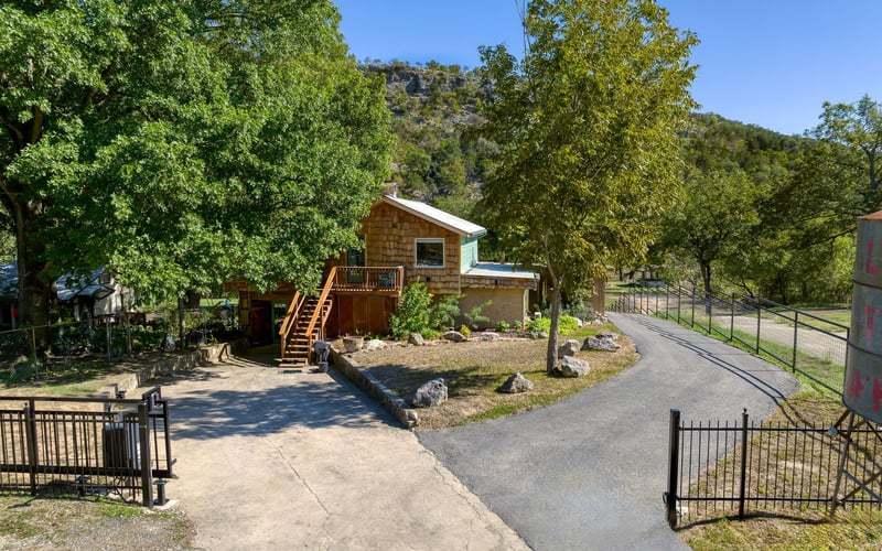 Rustic cabin-style rental home with wood siding and a wraparound deck, surrounded by lush trees and scenic hillside views, with a gated driveway entrance.