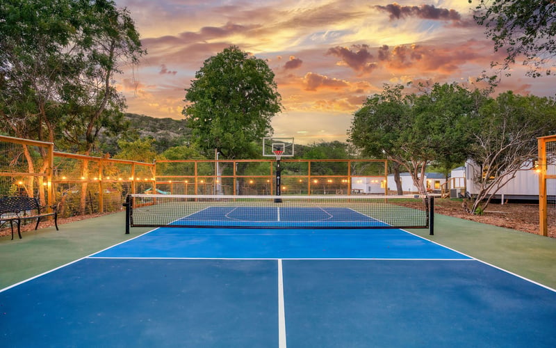 An outdoor pickleball and basketball court with a blue playing surface, surrounded by trees and string lights. The sky is illuminated with vibrant sunset colors, and small cabins are visible in the background.