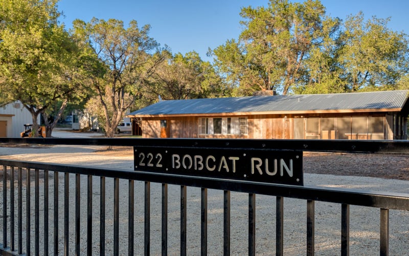 Black metal gate with a sign reading '222 Bobcat Run' in gold. A gravel driveway leads to a wooden building under tall trees.