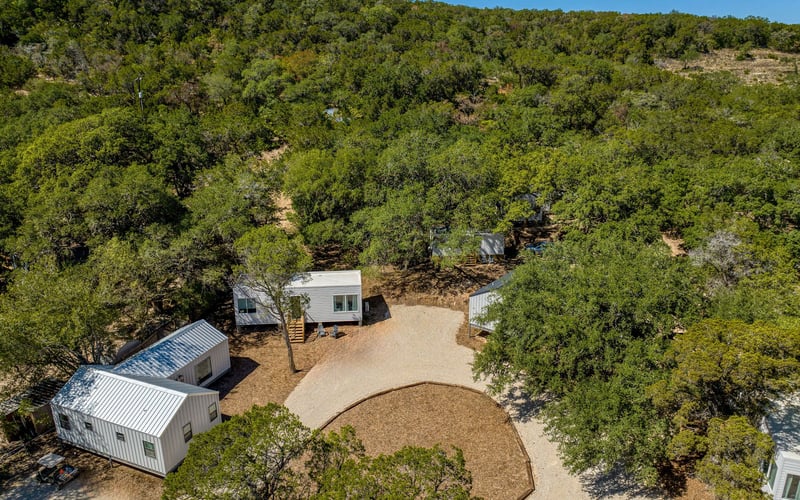 Aerial view of modern white tiny home rentals nestled among trees, featuring gravel pathways and a circular clearing, set in a peaceful hillside landscape.