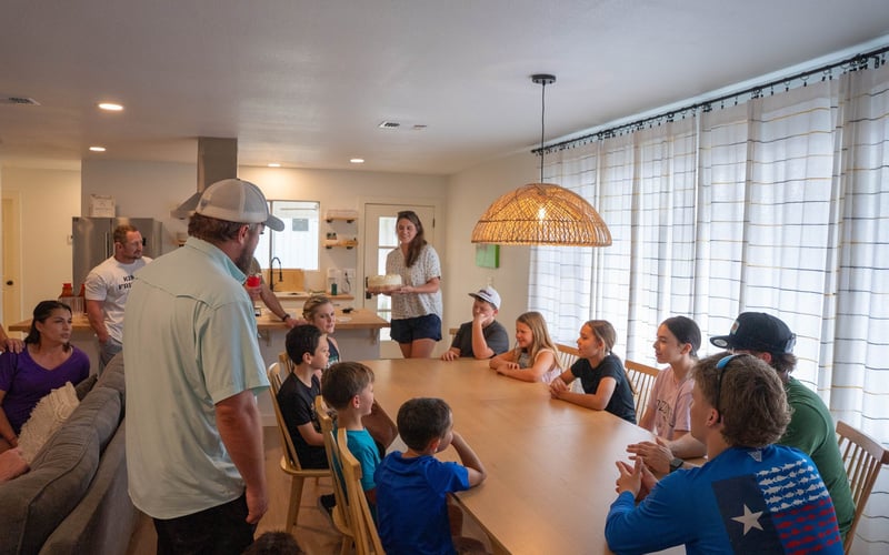 A group of children and adults gathered around a large wooden dining table in a bright kitchen, with one woman standing and holding a cake, possibly celebrating a birthday.