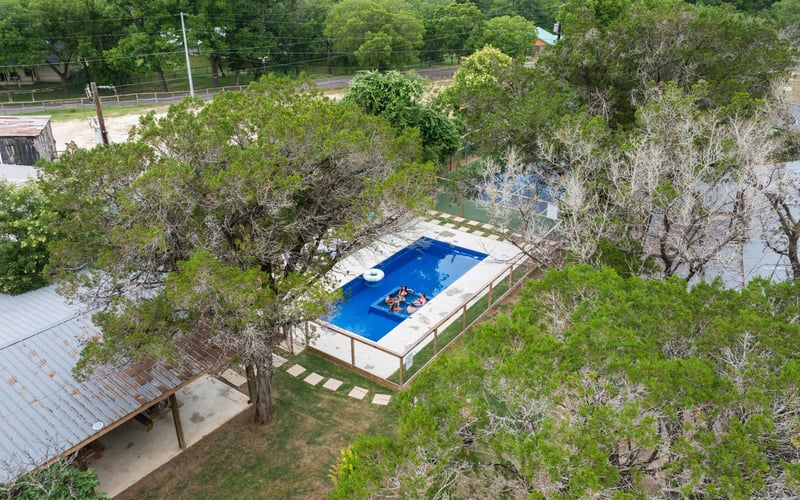 Aerial view of Texas Hill Country resort with a swimming pool surrounded by trees and fencing, with a few people swimming and relaxing in the pool.