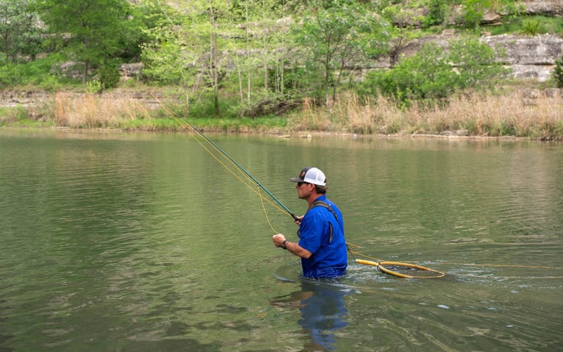 A man fishing in a river, standing in waist-deep water, surrounded by green trees and a rocky shoreline.