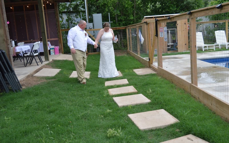 A bride and man walking on a pathway of large stepping stones in a backyard, holding hands. The bride is wearing a white wedding dress . The resort has a wooden fence, a pool area, and some outdoor furniture.