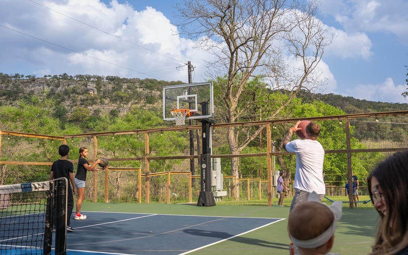 People playing basketball outdoors on a court with a backdrop of green trees and mountains under a partly cloudy sky.